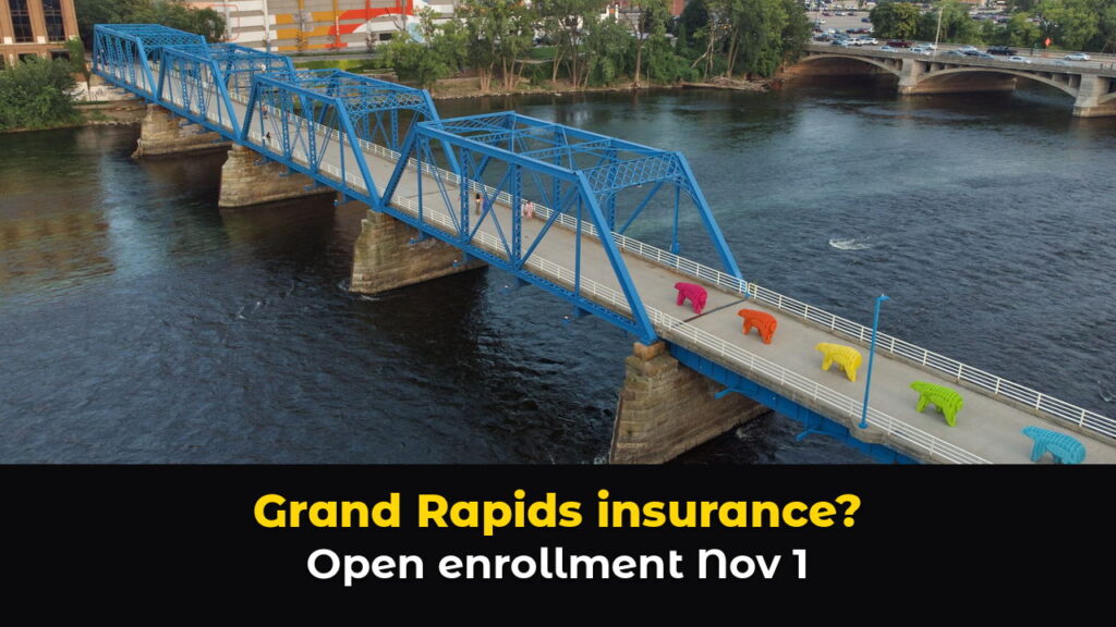 Aerial view of the Blue Bridge footbridge over the Grand River in downtown Grand Rapids, Michigan, with colorful bear sculptures on the deck, above hook text: Grand Rapids insurance? Open enrollment Nov 1.